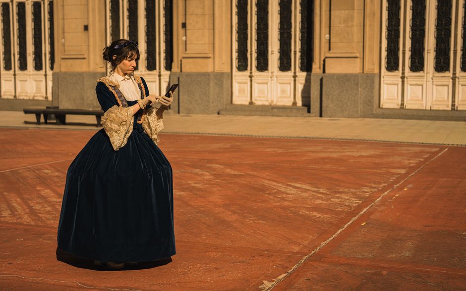 A woman in Victorian attire using a smartphone outside a historic building.
