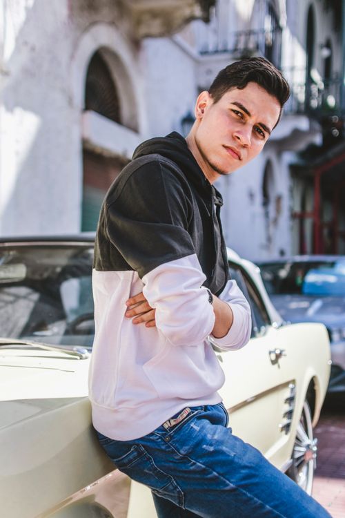 Free Young man in casual attire leaning on a classic car outdoors with urban backdrop. Stock Photo