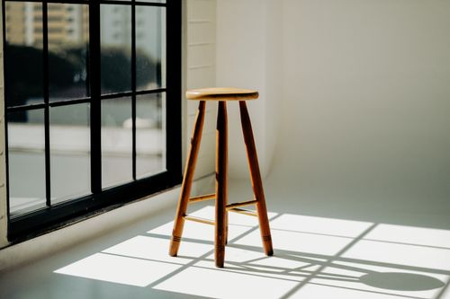 Free A minimalistic scene with a wooden stool bathed in sunlight next to a window. Stock Photo