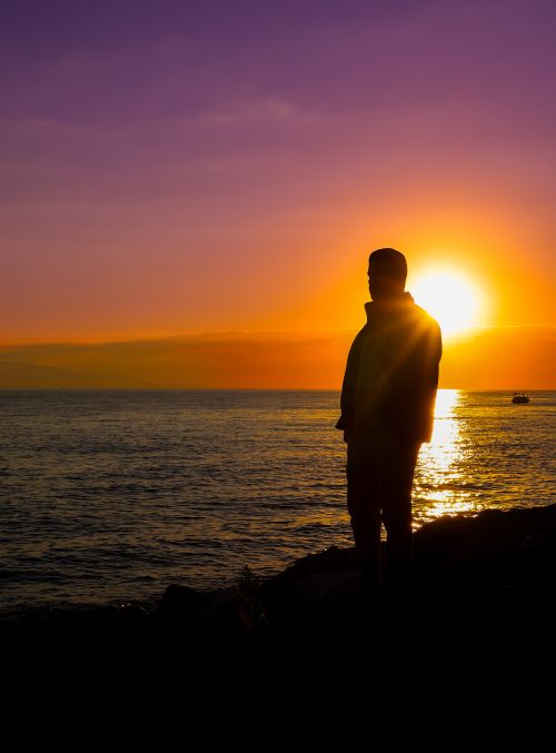 Free A solitary figure stands by the sea, silhouetted against a vibrant sunset, capturing a moment of contemplation and serenity. Stock Photo