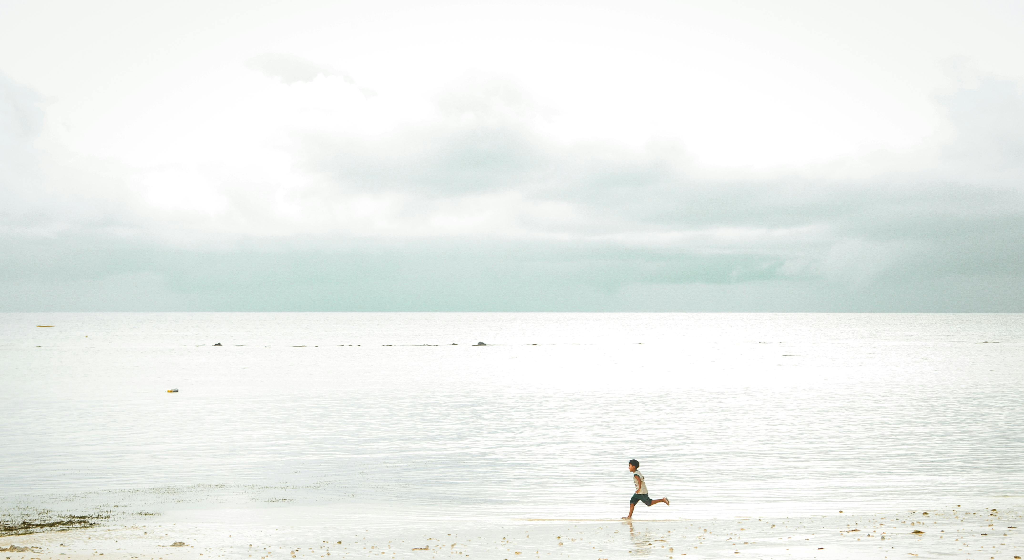 A boy runs joyfully along a serene seaside, under a cloudy sky.