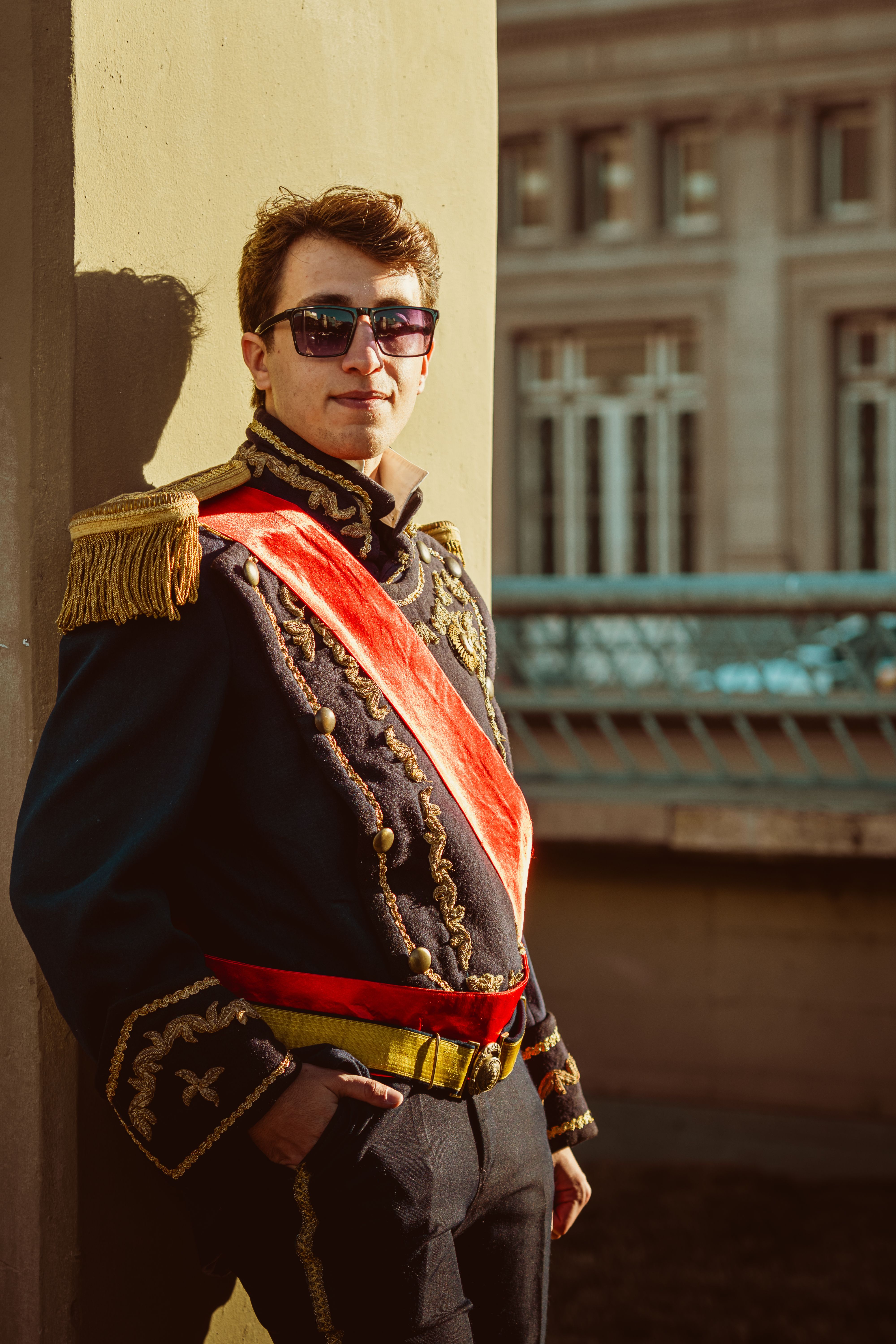 Stylish man in regal uniform and sunglasses posing confidently outdoors. Distinctive urban architecture.