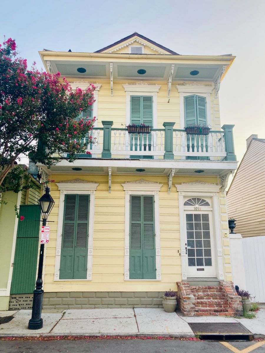 A yellow two-story New Orleans house with green shutters, an iron balcony and pink crape myrtle
