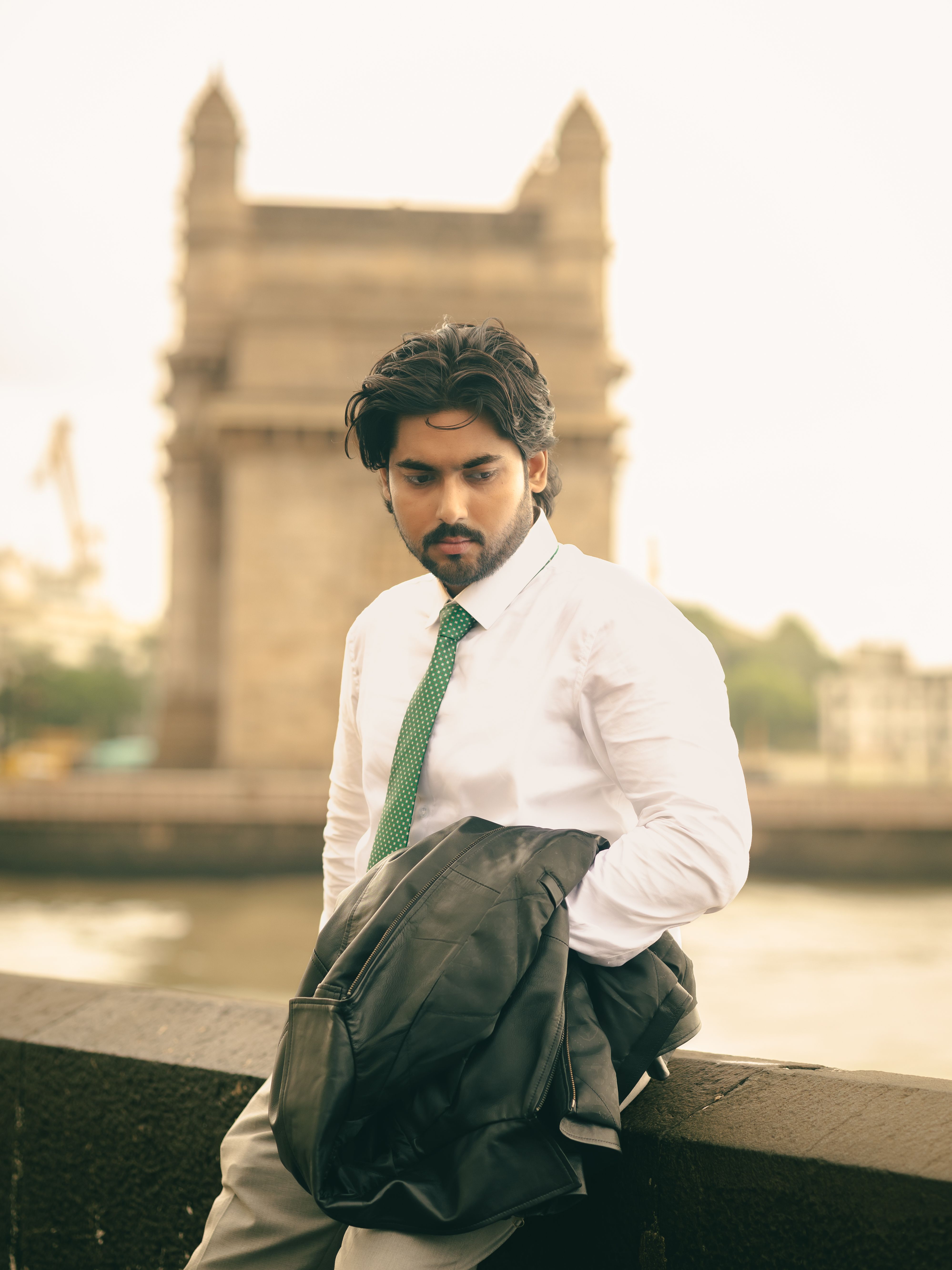 A fashionable man poses with the Gateway of India in the background, wearing a white shirt and holding a jacket.