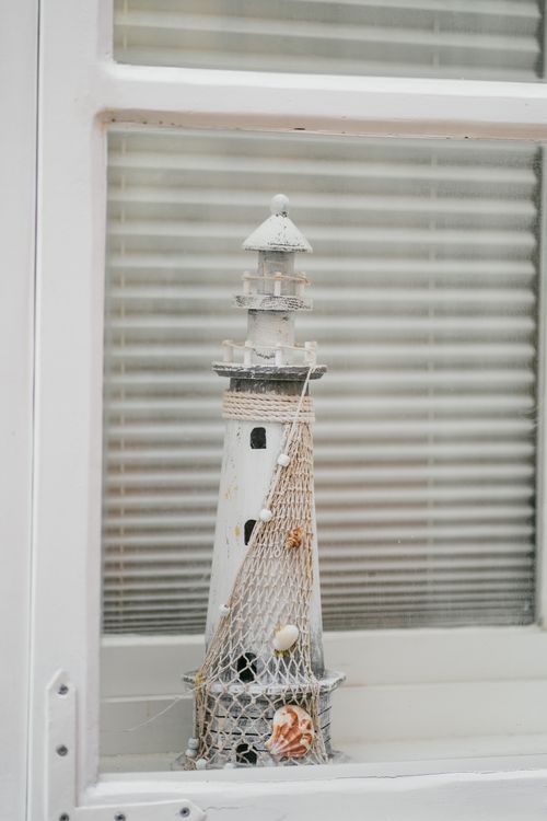Free White decorative lighthouse figurine against window blinds, creating a nautical theme indoors. Stock Photo