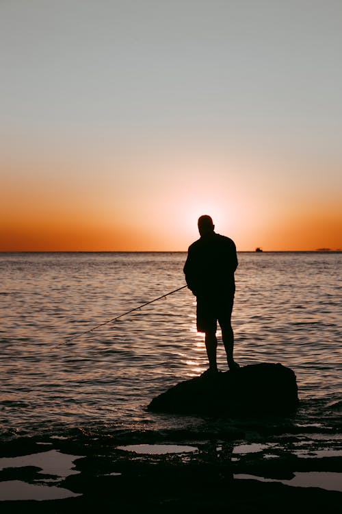 Silhouette Of Man Holding Fishing Rod Standing On A Rock During Sunset