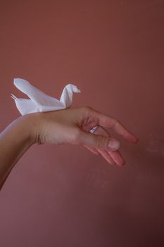 Close-up of a hand holding a white origami crane against a muted pink wall.