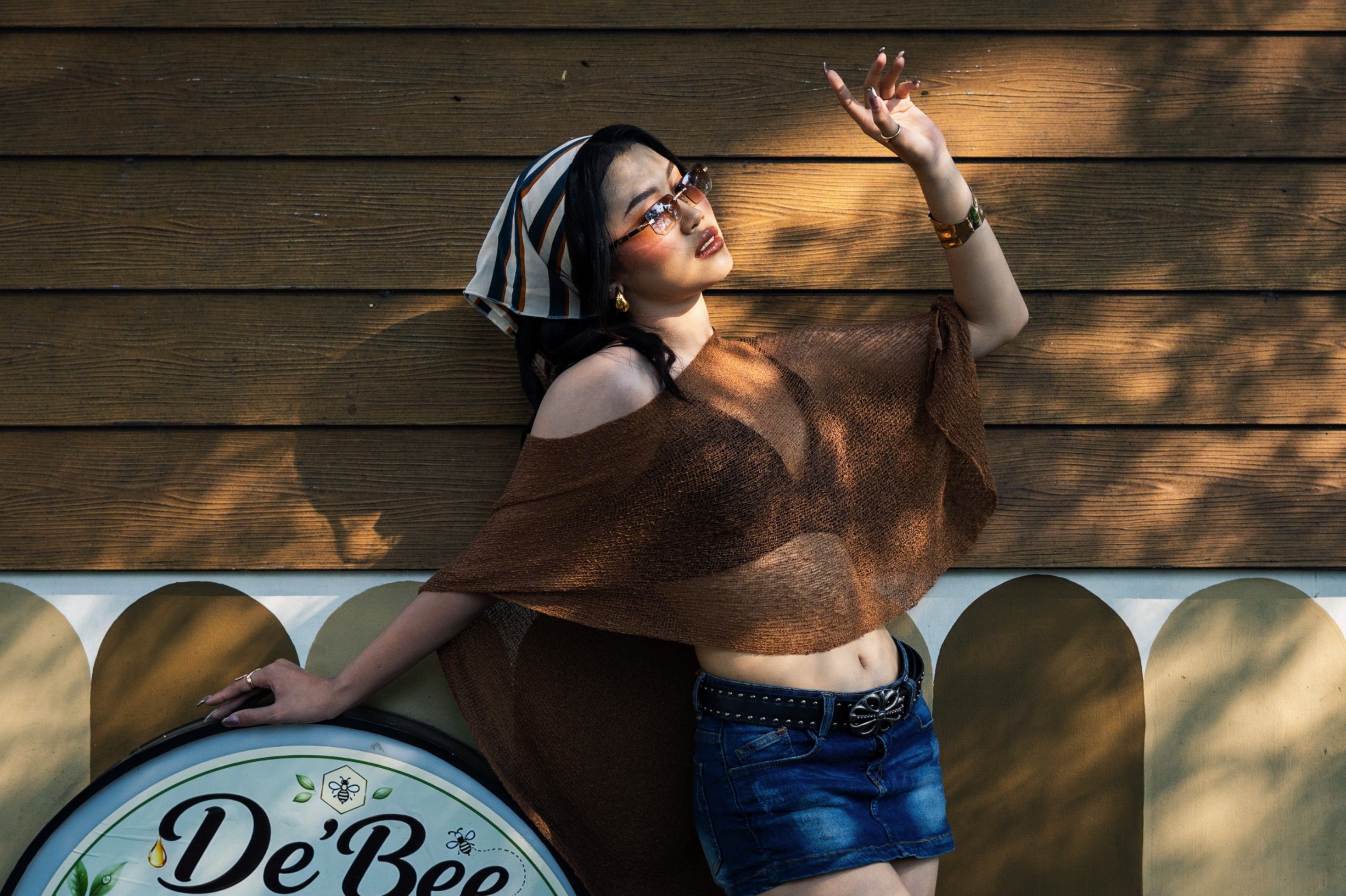 Fashionable woman in sunglasses and headscarf poses outdoors against a wooden wall in Jakarta.