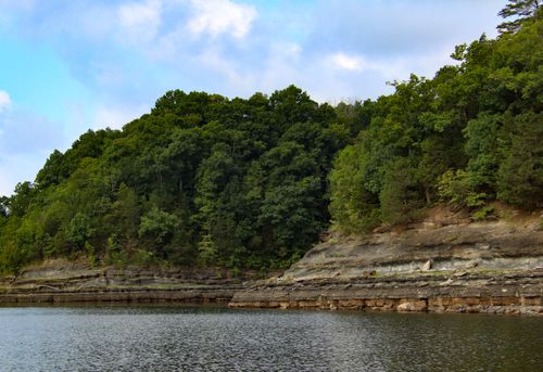 Free A lush green forest atop a rocky cliffside beside a serene lake under a blue sky. Stock Photo