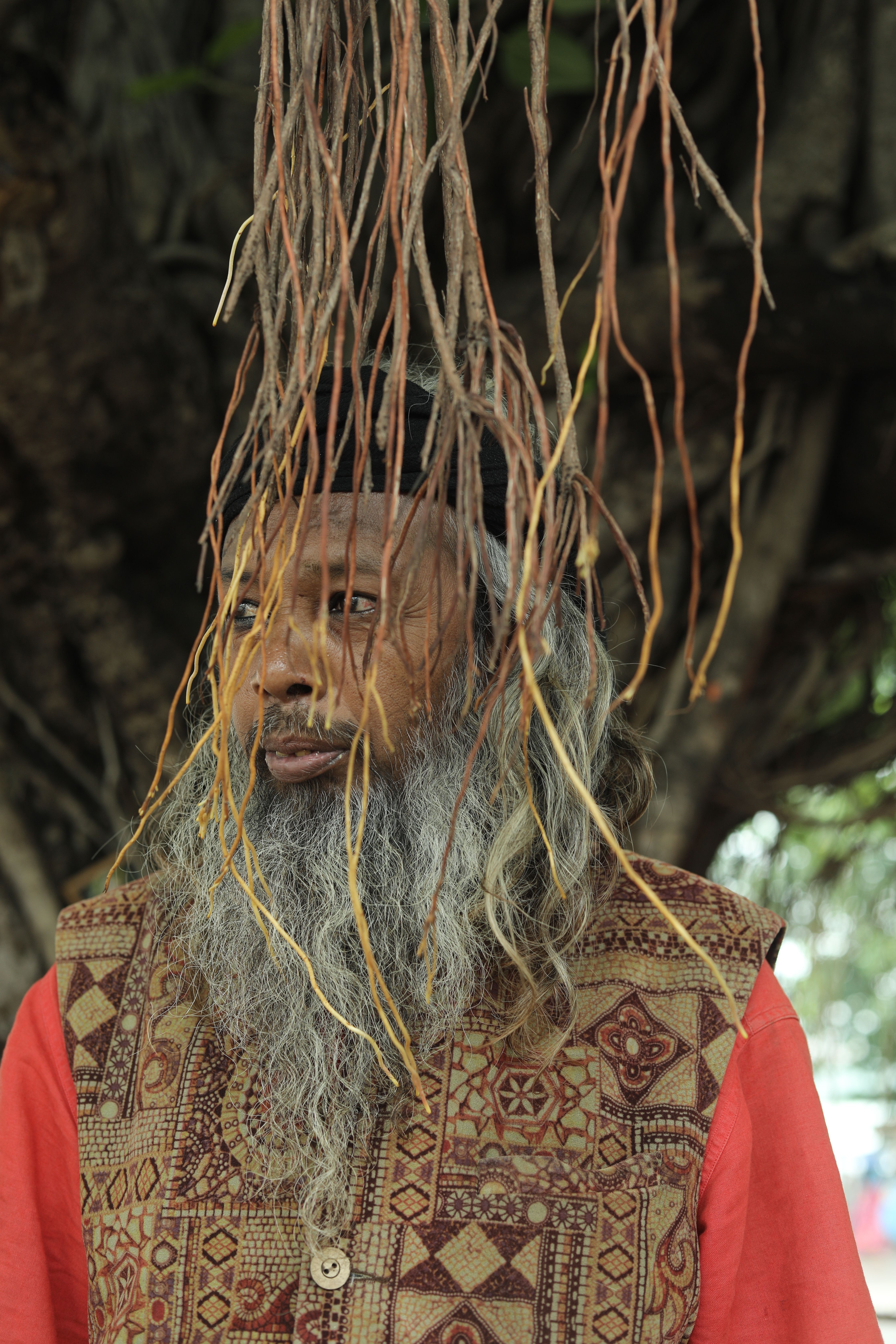 A man with a long beard stands under banyan roots in Raipur, India.