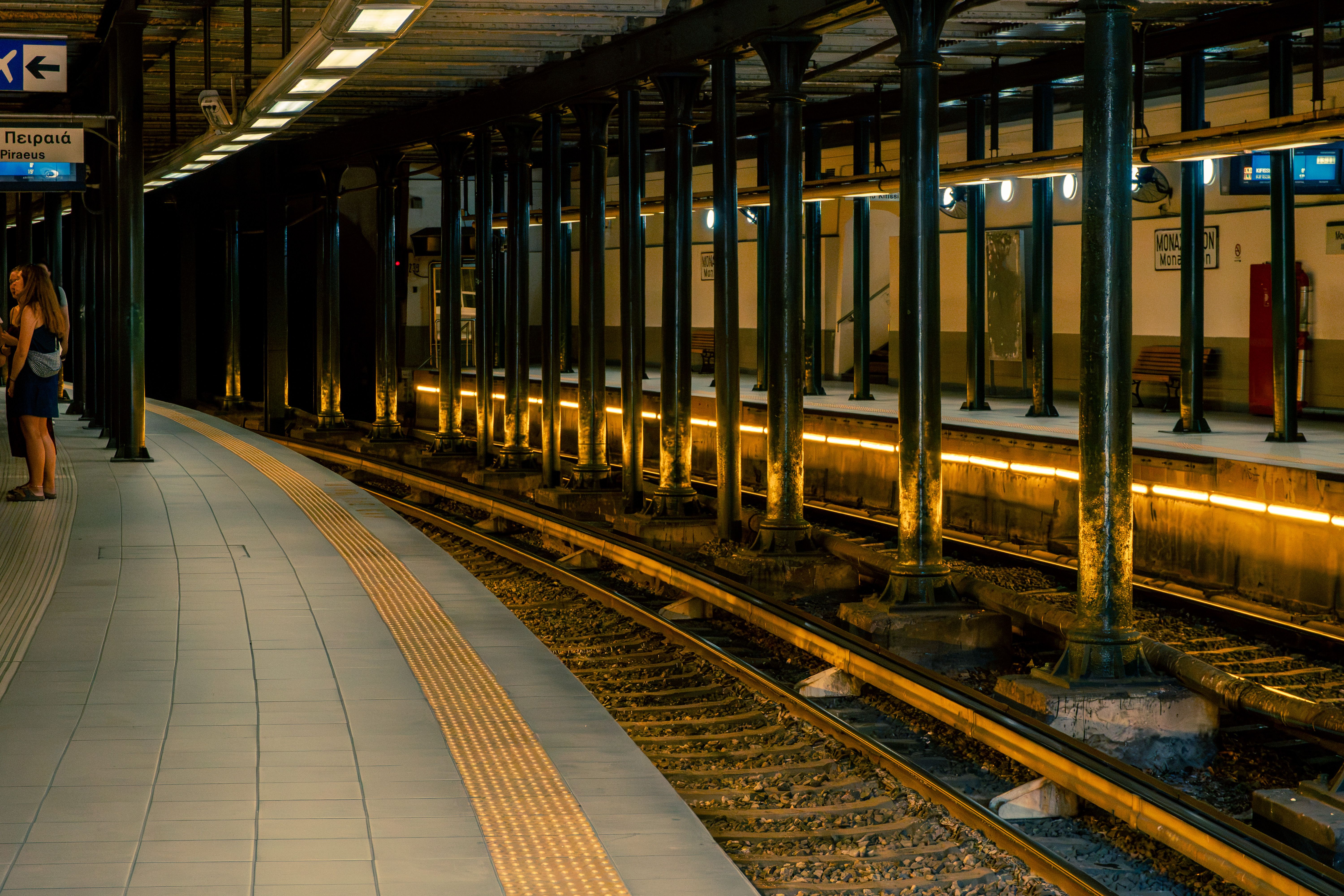 Moody nighttime scene of Athens subway platform with yellow lights illuminating empty tracks and few passengers.