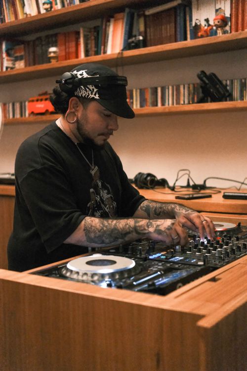 Free A tattooed DJ mixes music with a console in a cozy indoor setting with bookshelves in the background. Stock Photo