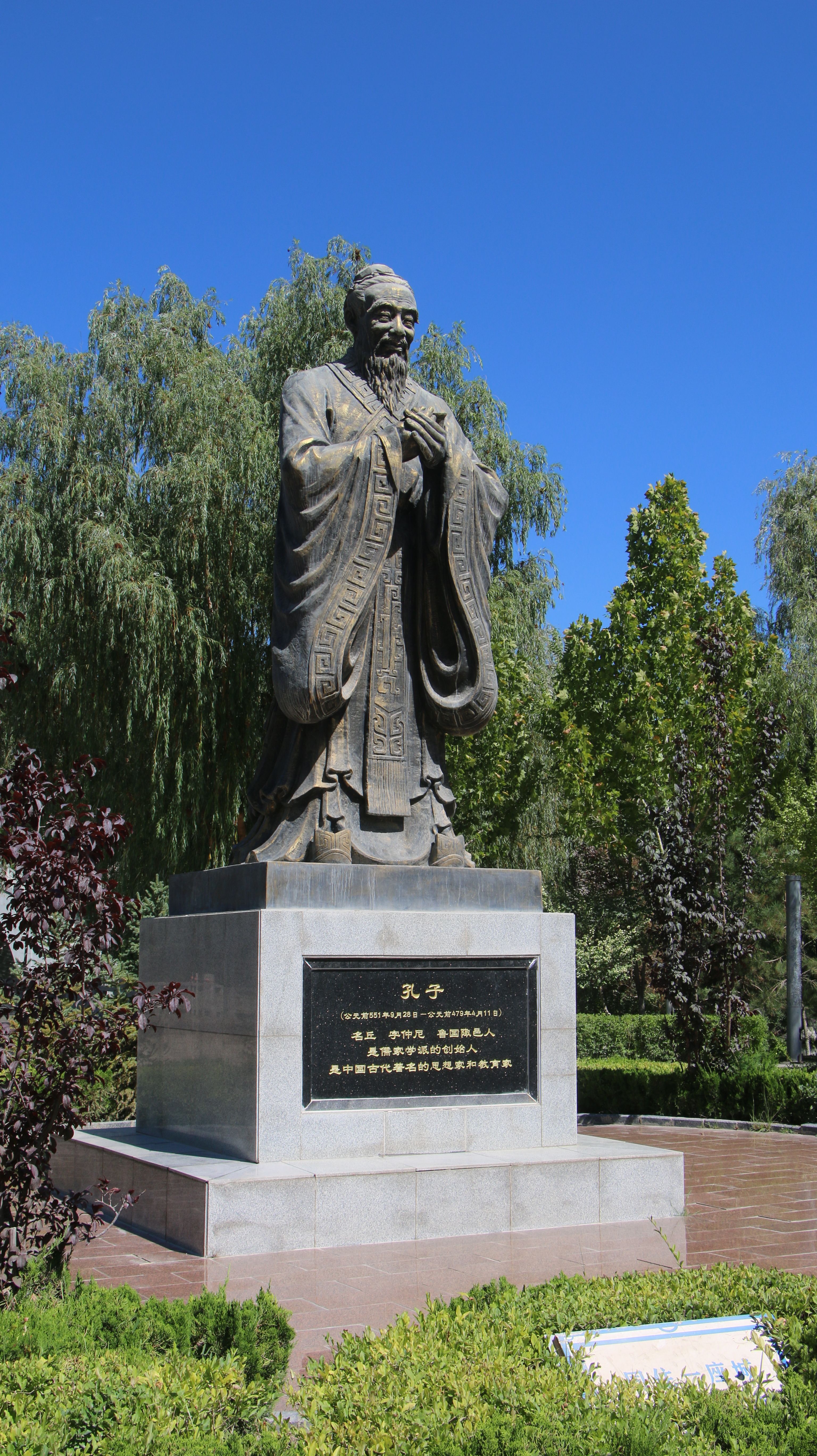 Bronze statue of Confucius surrounded by greenery under a clear sky.