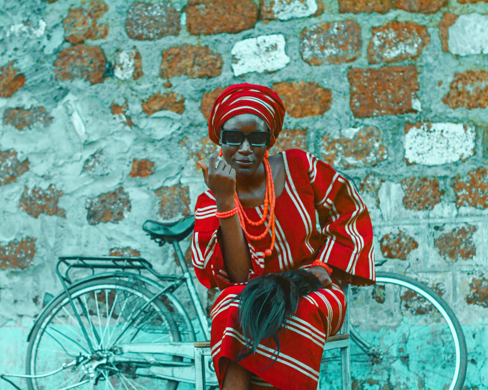 African woman in vibrant traditional outfit, seated by a bicycle against rustic wall.