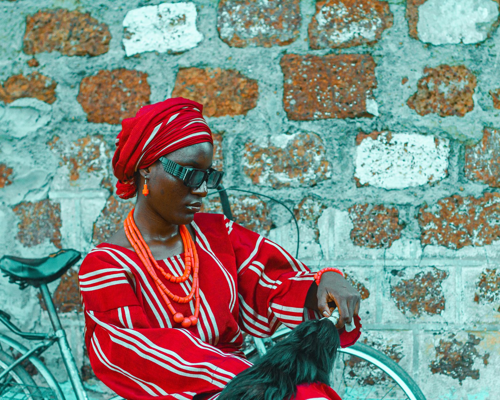 A Nigerian woman in vibrant traditional attire sits by a rustic stone wall with a bicycle.