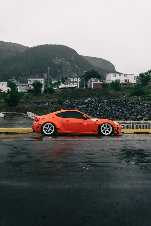 Free A vibrant orange sports car parked on a wet coastal road with scenic hills in the background. Stock Photo