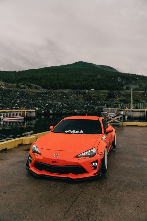 Free A vivid orange sports car parked by a waterfront with lush green hills in the background. Stock Photo
