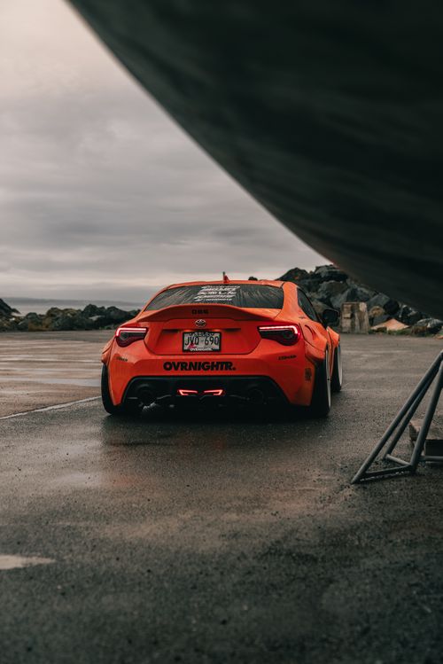 Free Capture of a vibrant orange sports car parked on a wet pavement under an overcast sky. Stock Photo
