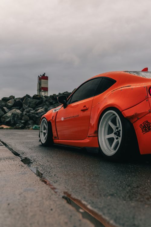 Free Dynamic shot of a sleek orange sports car on a wet road next to coastal rocks under a cloudy sky. Stock Photo
