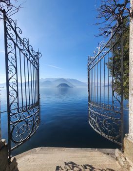Discover a serene lake view framed by elegant open iron gates under a clear blue sky.