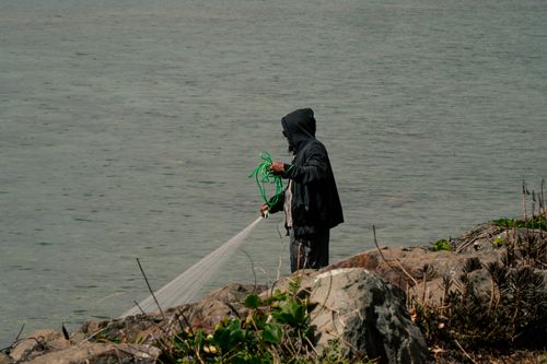 Free A fisherman in a hoodie casts a net into the calm waters by the rocky shoreline. Stock Photo