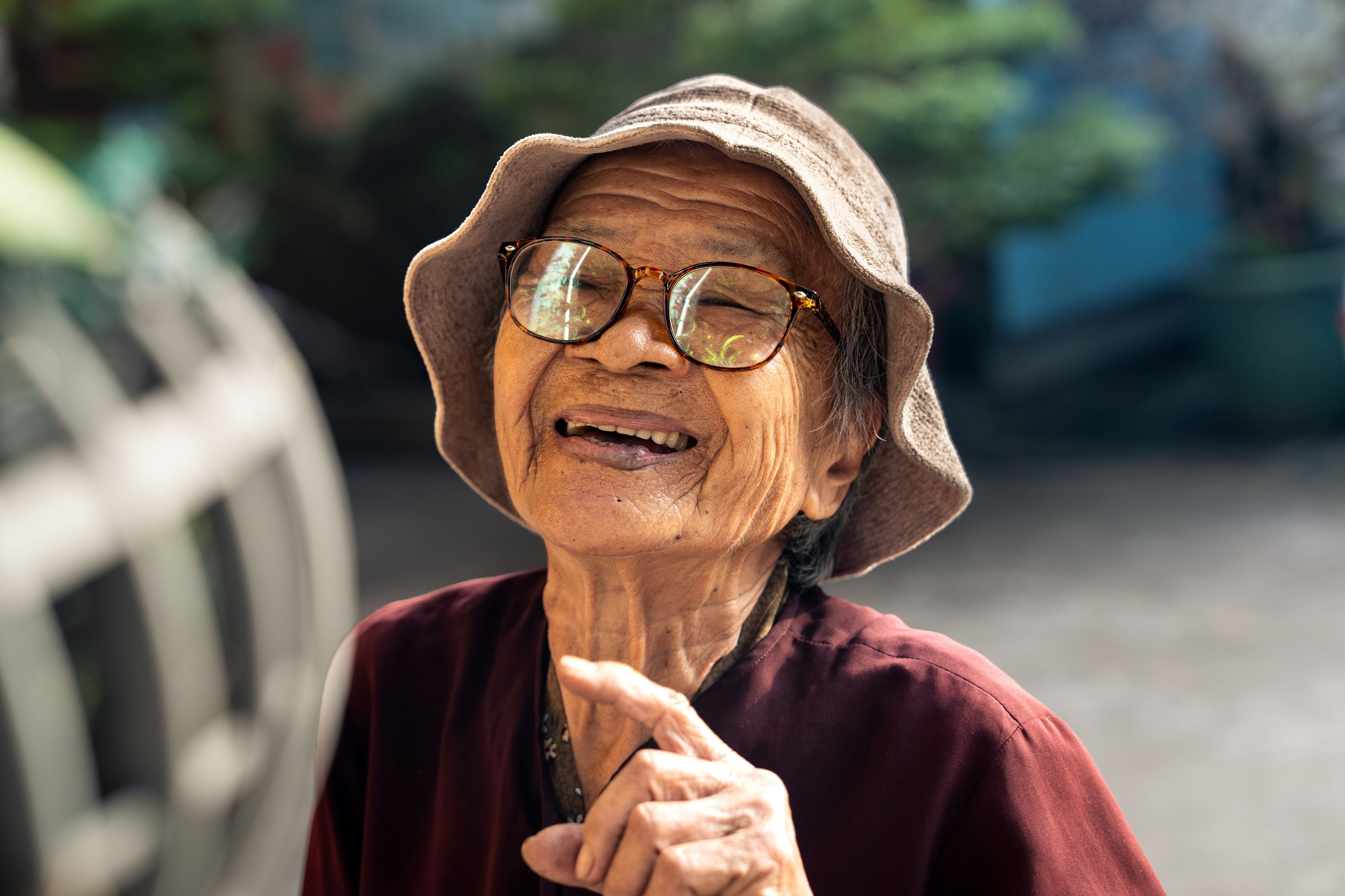 Elderly Vietnamese woman smiling joyfully outdoors in sunlight, wearing glasses and a sunhat.