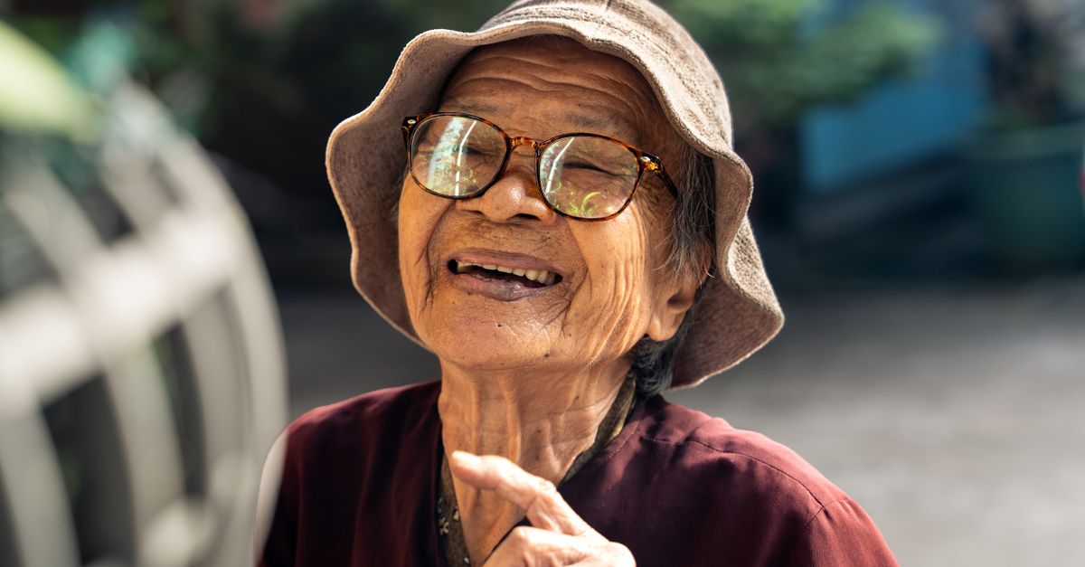 Elderly Vietnamese woman smiling joyfully outdoors in sunlight, wearing glasses and a sunhat.