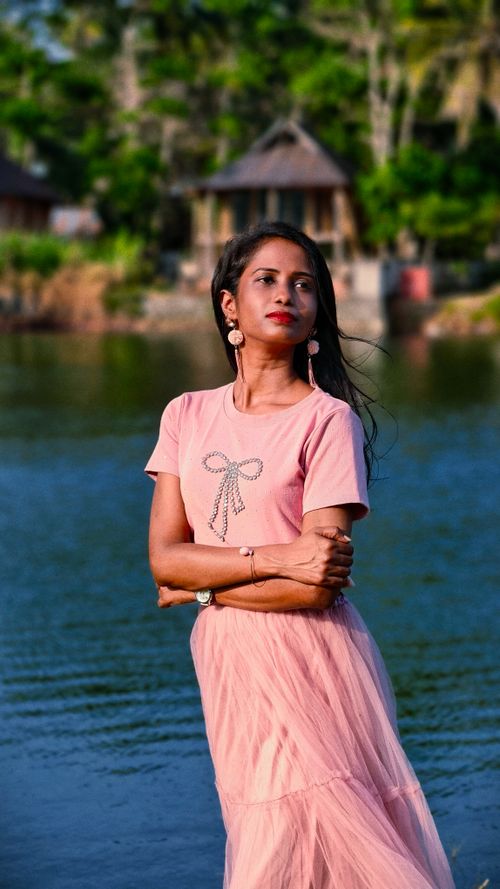 Free Woman in pink dress poses by serene lake with lush greenery in background. Stock Photo