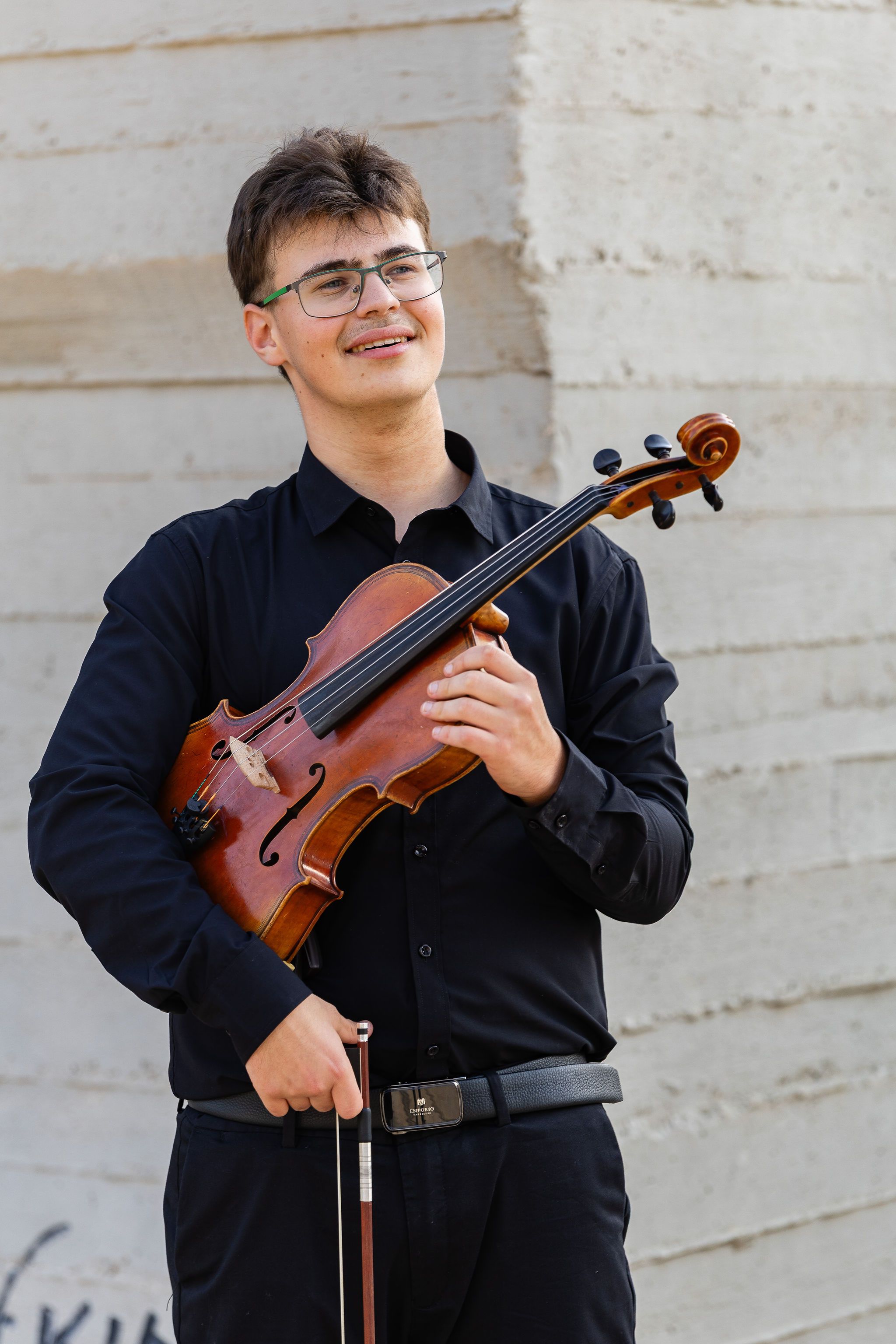 Young male violinist in black attire holding a violin outdoors.