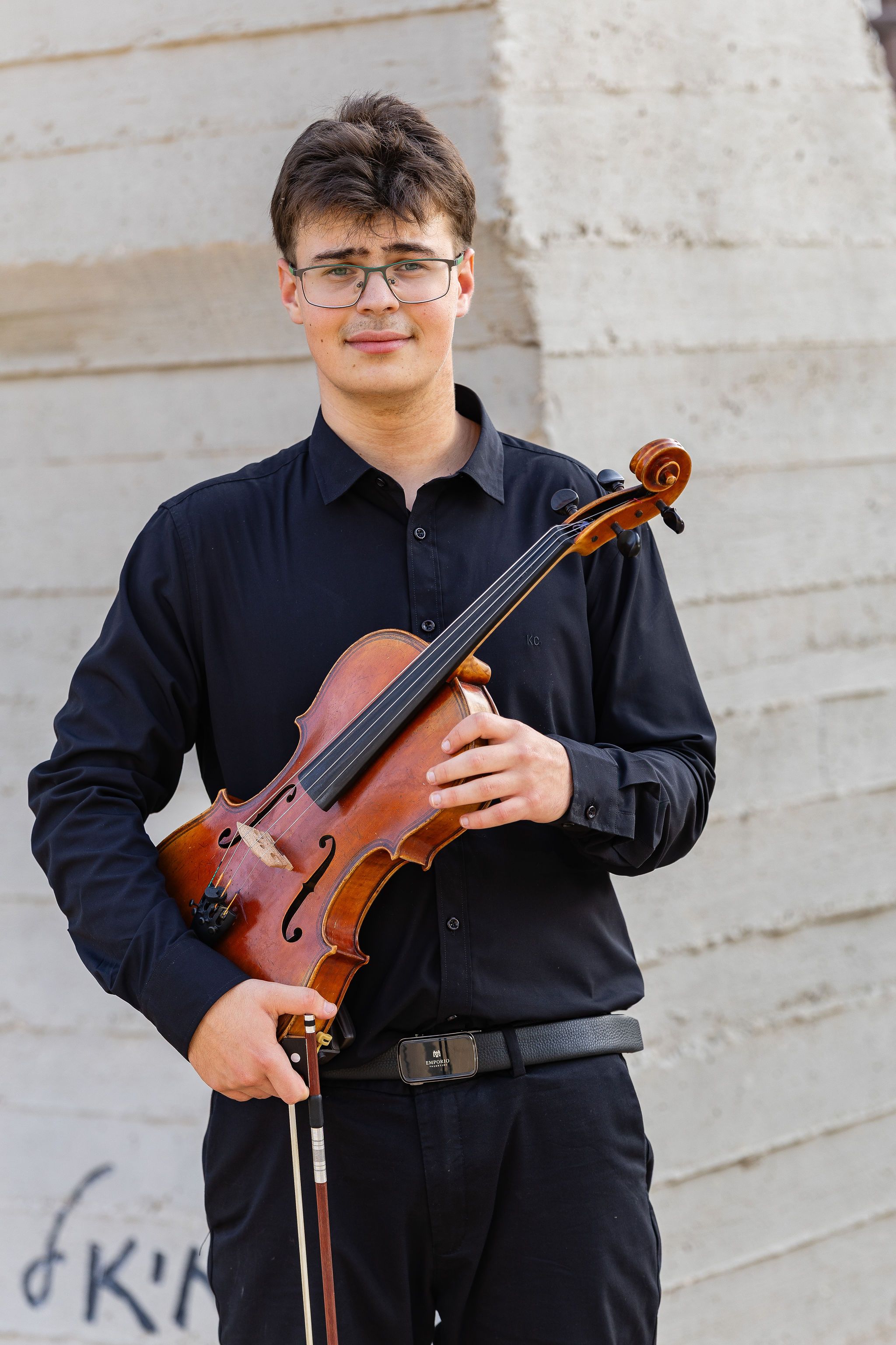 Smiling young musician in black holding a violin outdoors against a concrete wall.