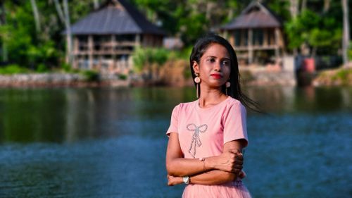 Free A woman in a pastel pink outfit poses by a tranquil lakeside resort during summer. Stock Photo