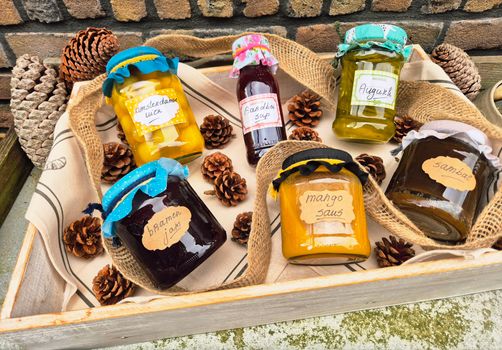 A collection of colorful homemade jams and sauces in jars with decorative lids displayed with pinecones.
