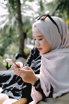 A young woman in a hijab enjoys outdoor leisure time, browsing her phone in a relaxed setting.