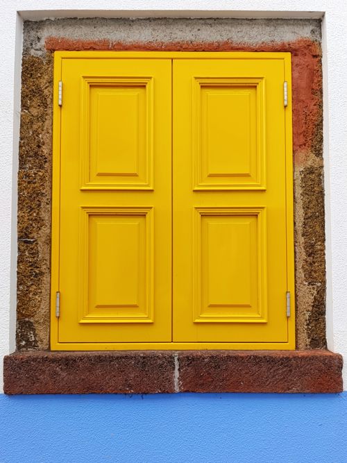 Free A yellow shuttered window contrasts with a textured stone frame and blue wall. Stock Photo