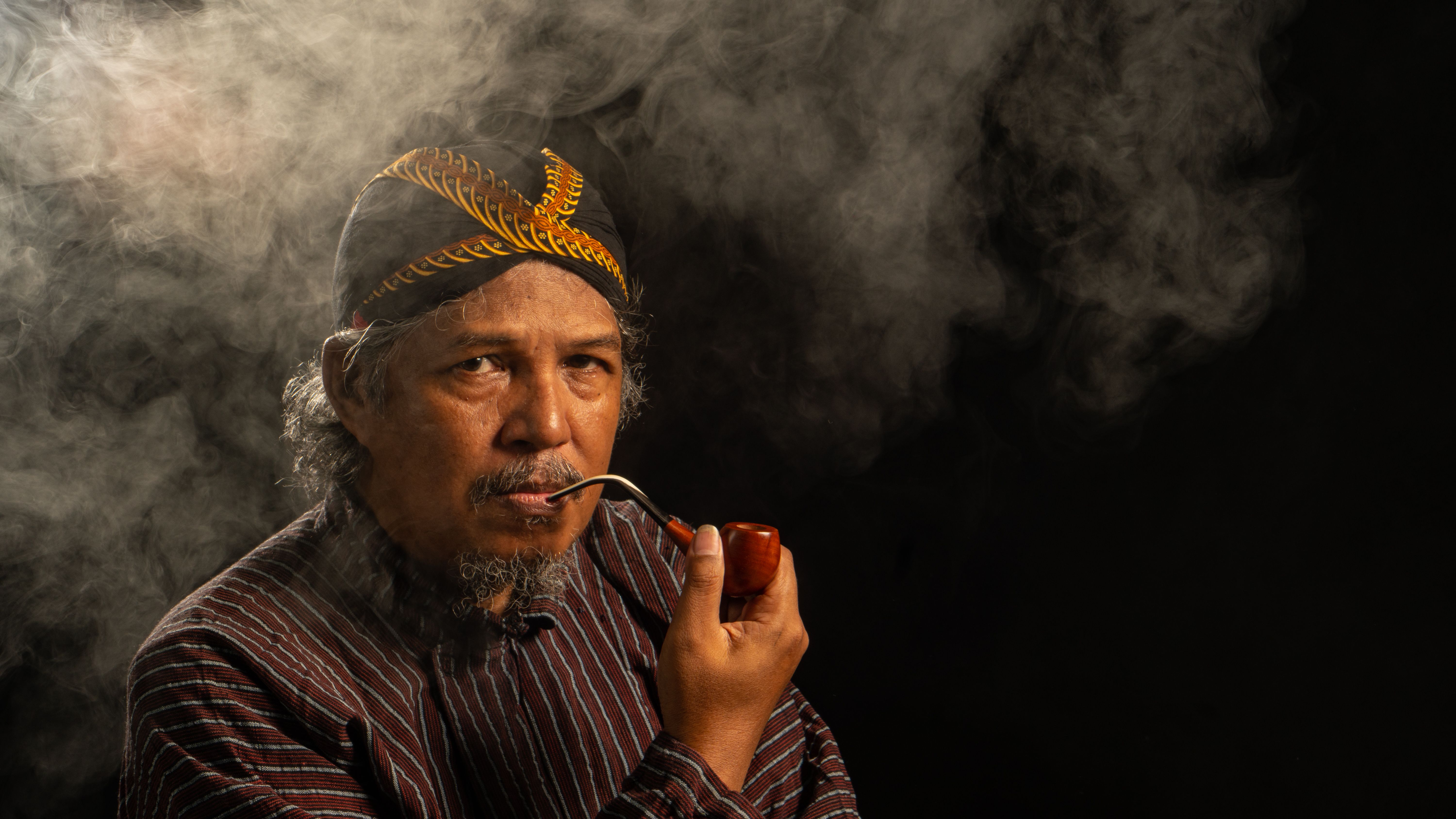 Low-key portrait of a senior Javanese man smoking a pipe with dramatic smoke effects.