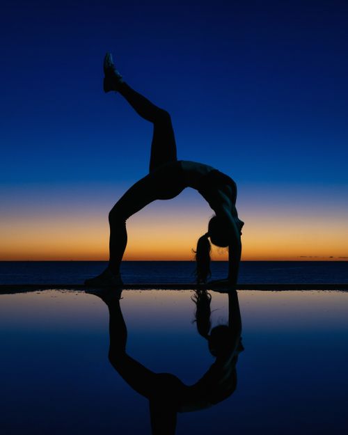 Free A person practices a yoga pose at sunset, reflecting on water with a vibrant sky. Stock Photo