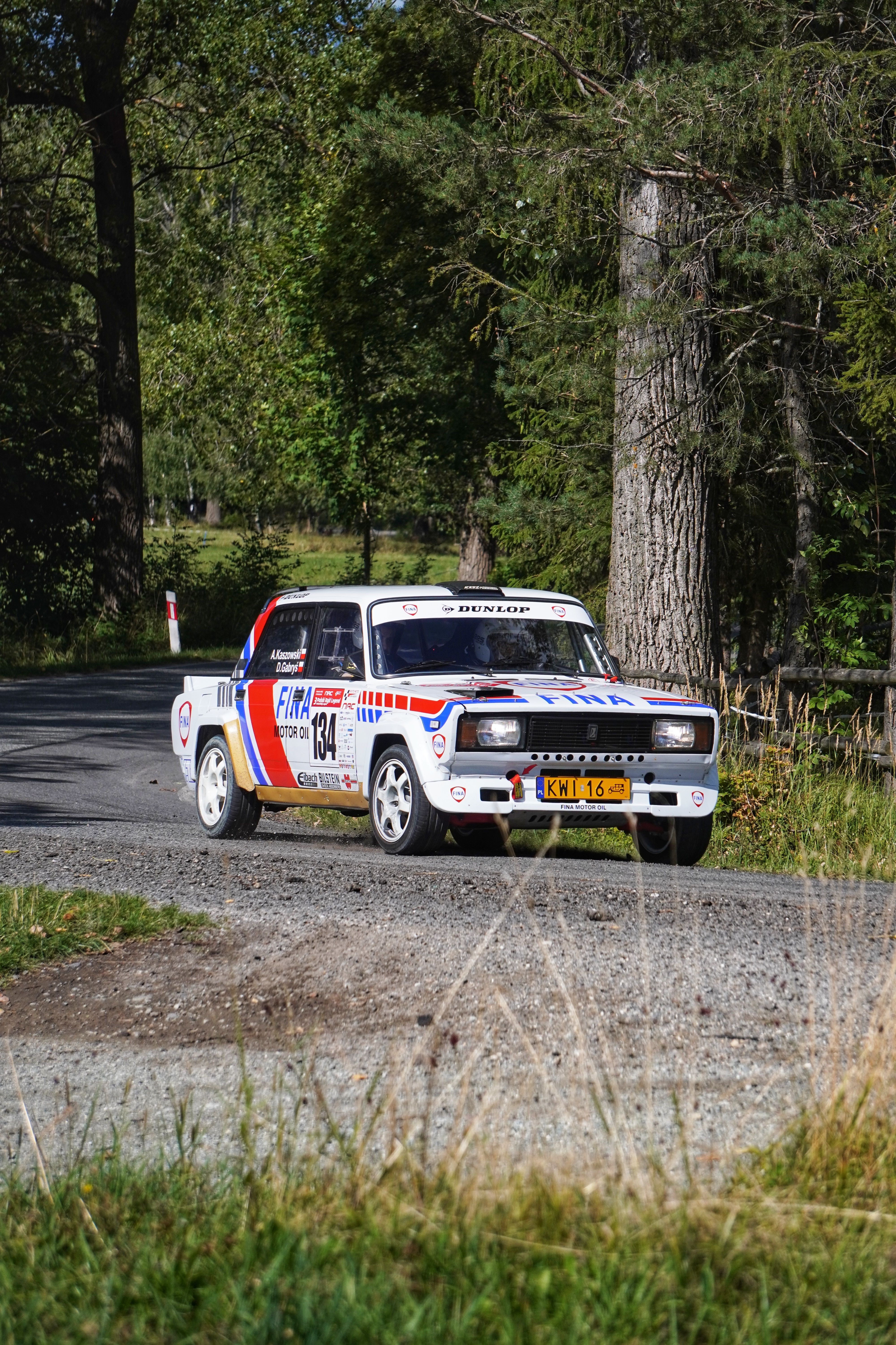 A vintage rally car drives along a picturesque forest road in bright daylight, showcasing motorsport action.