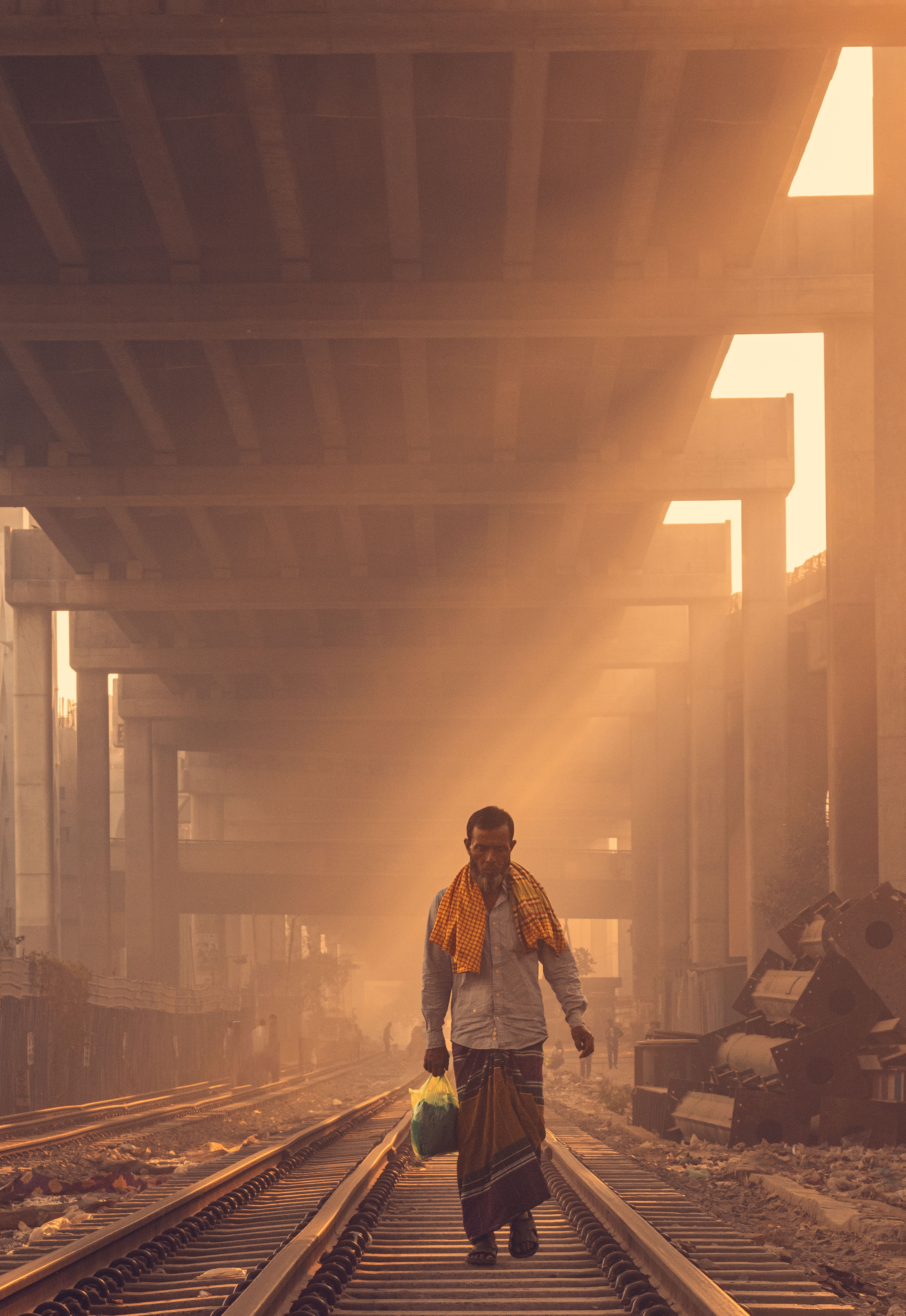 A man walks on railway tracks beneath an overpass at sunrise, creating a moody scene.