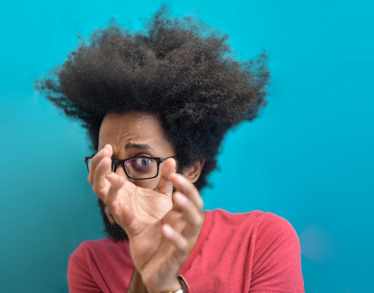 Scared Young Ethnic Man Looking Through Hands On Blue Background In Studio