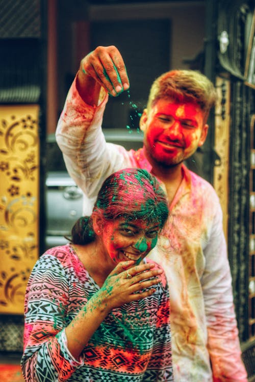 Free Positive man spilling colorful powder on laughing Indian woman while having fun during Holi festival on town street Stock Photo