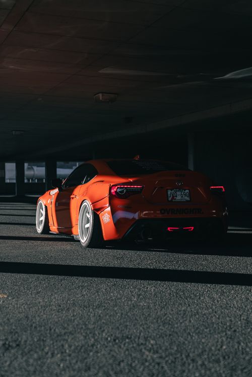 Free Low-angle view of an orange sports car parked in a dimly lit urban garage, showcasing sleek design and vibrant color. Stock Photo