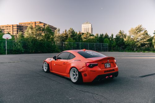 Free A sleek orange sports car parked outdoors with urban buildings and trees in the background. Stock Photo