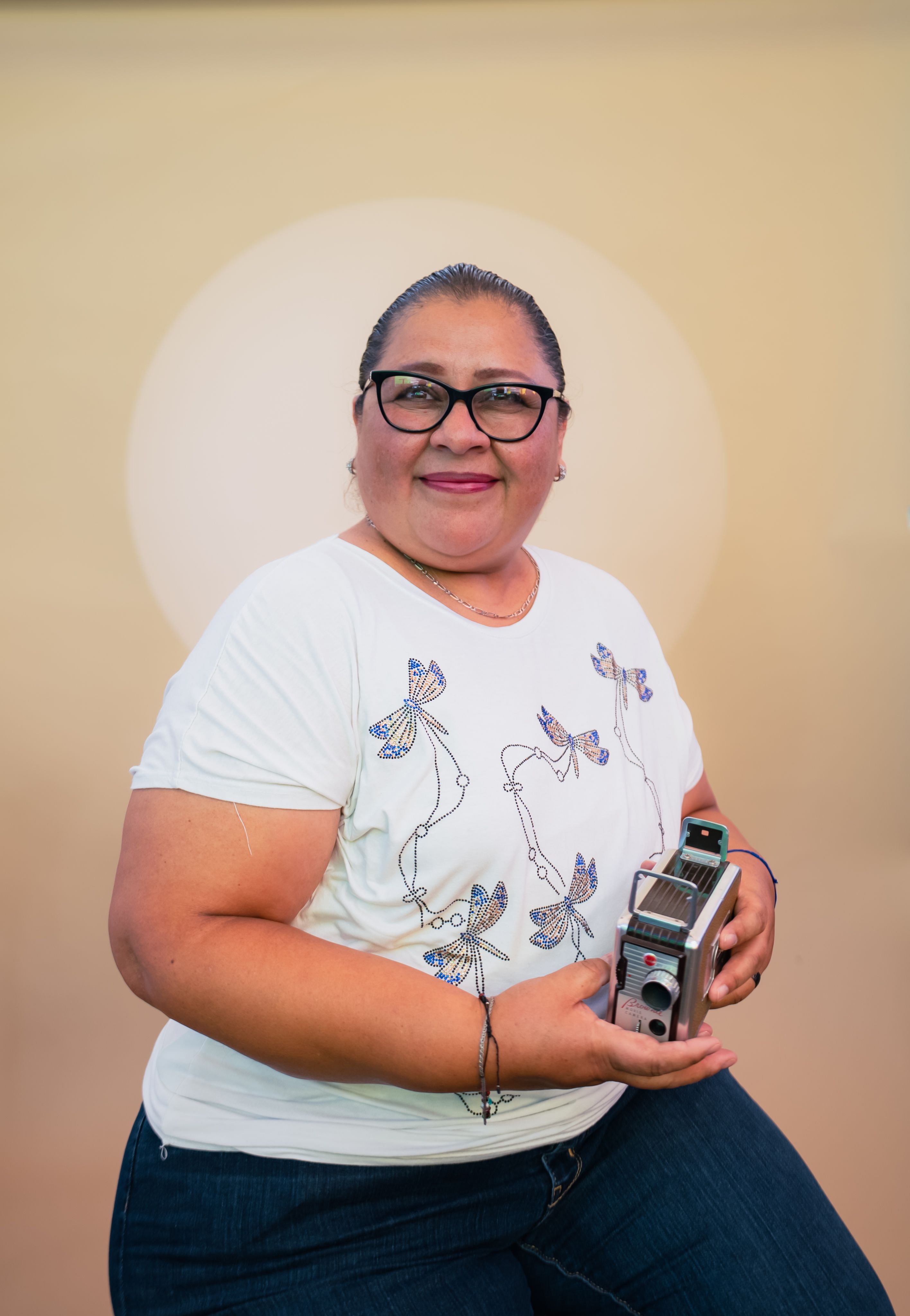 Smiling woman in glasses holds vintage camera against a soft yellow background.
