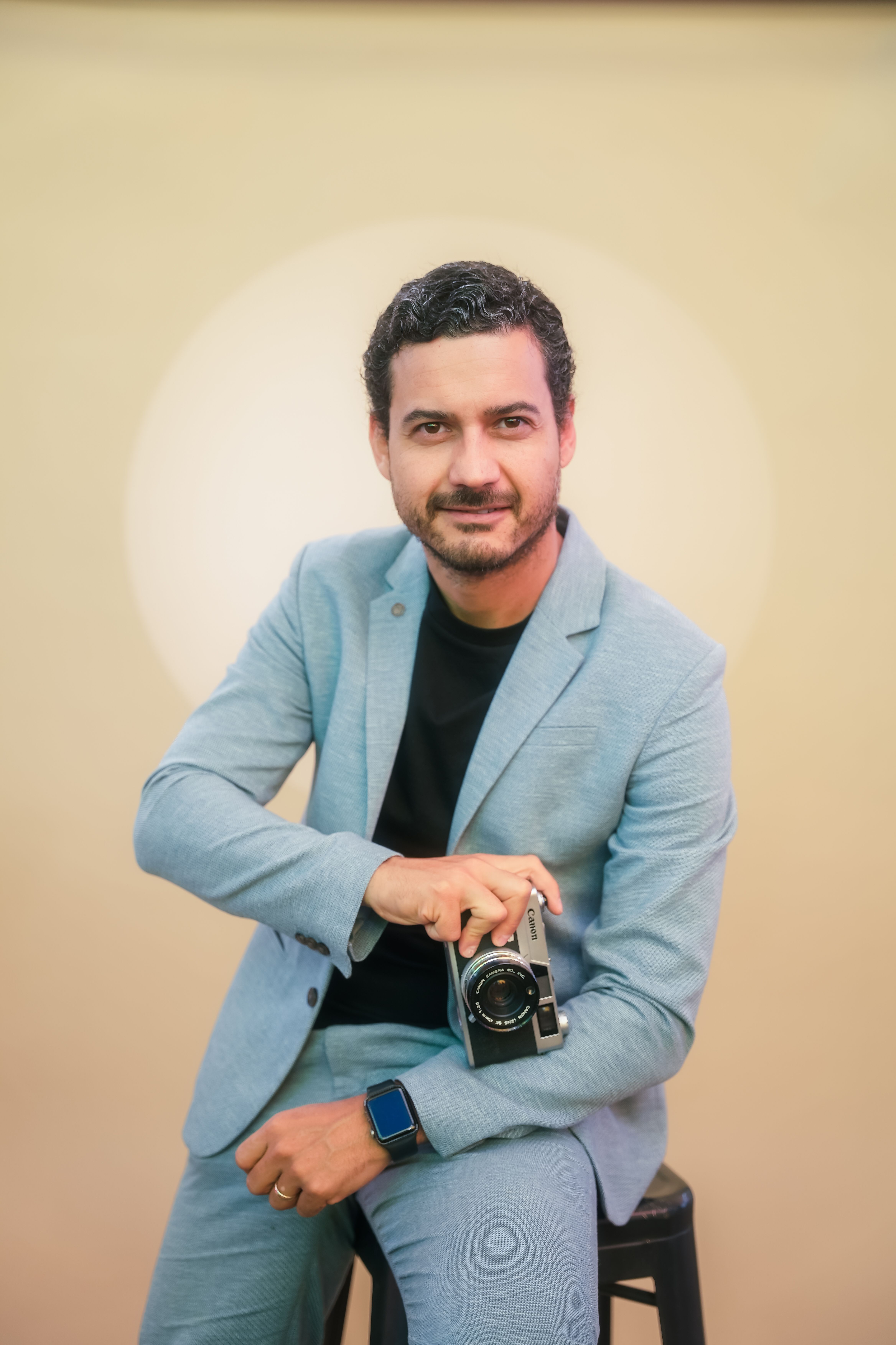 A man in a blue suit holds a camera, seated in a studio with soft lighting.