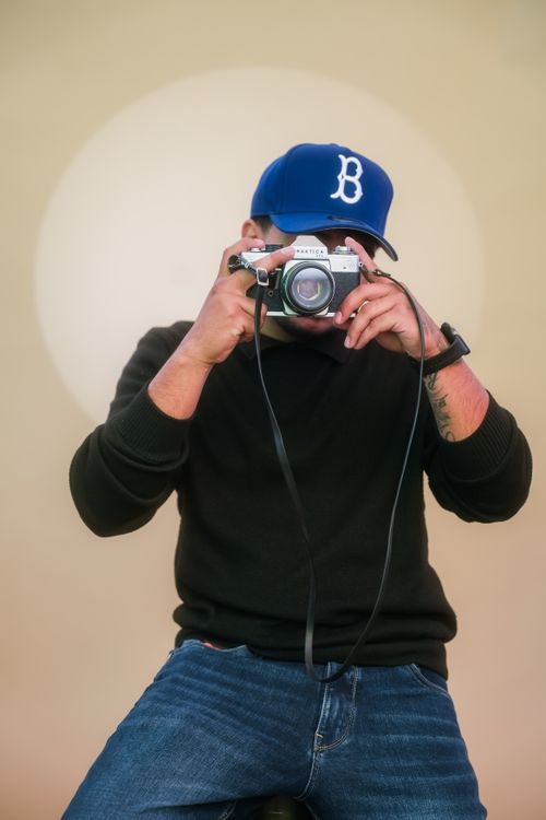 Free Portrait of a man in a blue cap using a vintage analog camera, seated against a neutral background. Stock Photo