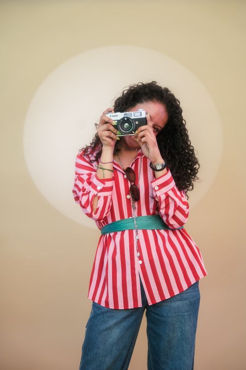 Free Young woman in a striped shirt holding a camera against a circular light backdrop, capturing a joyful moment. Stock Photo
