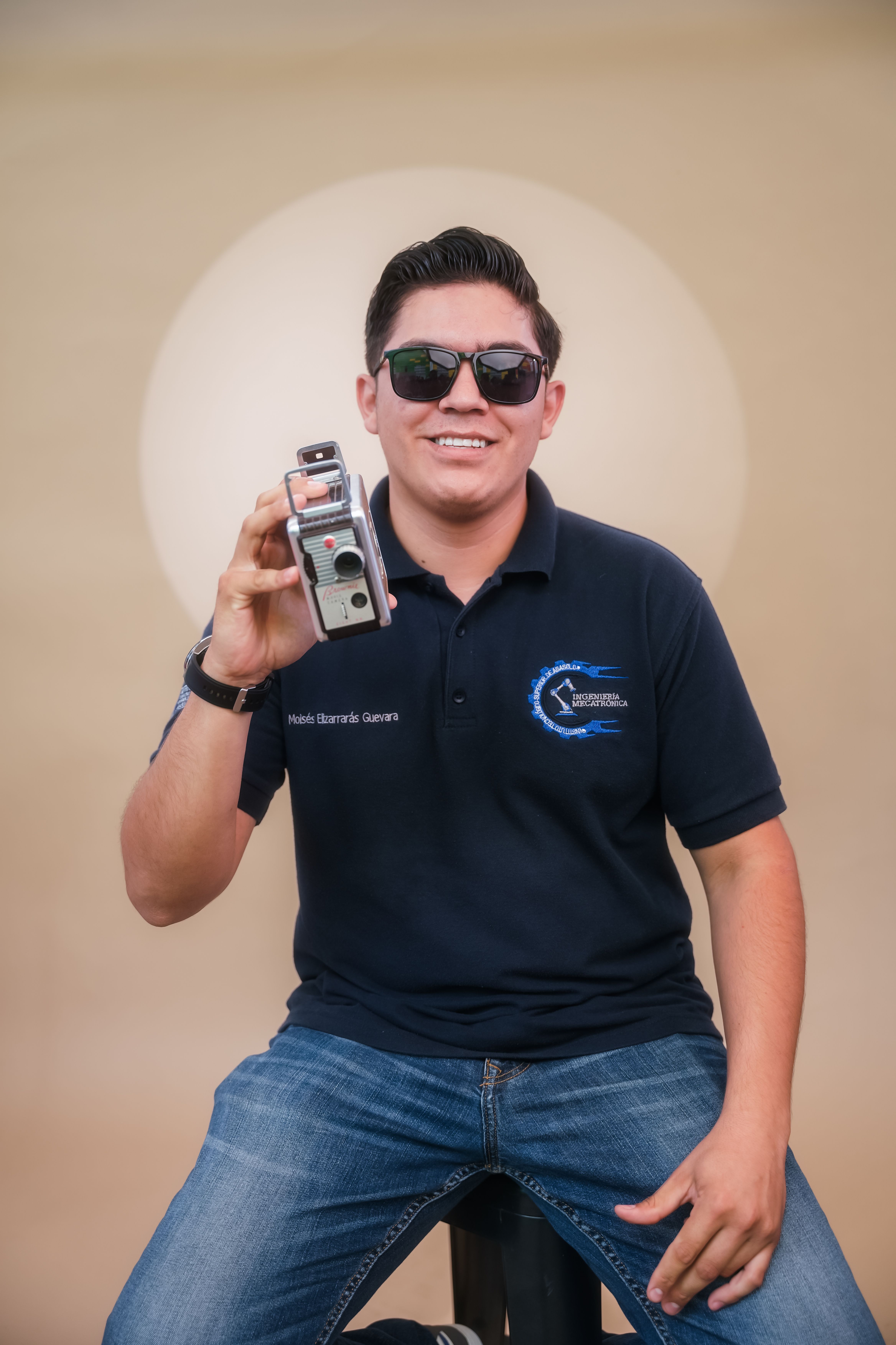 Smiling young man wearing sunglasses, holding a vintage camera against a soft background. Captured outdoors in daylight.