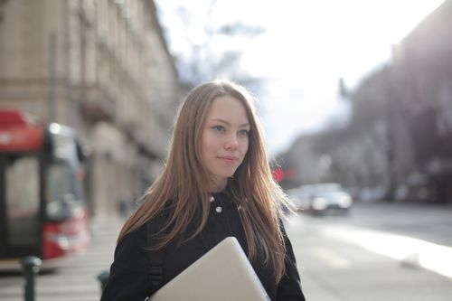 Free Confident young woman holding a laptop while walking on a bustling city street. Stock Photo