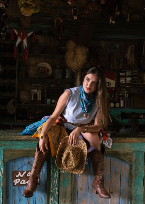 Free Portrait of a young cowgirl sitting on a rustic bar holding a hat indoors. Stock Photo