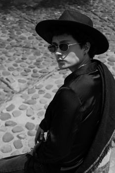 Black and white photo of a man in sunglasses and hat sitting on a cobblestone street.